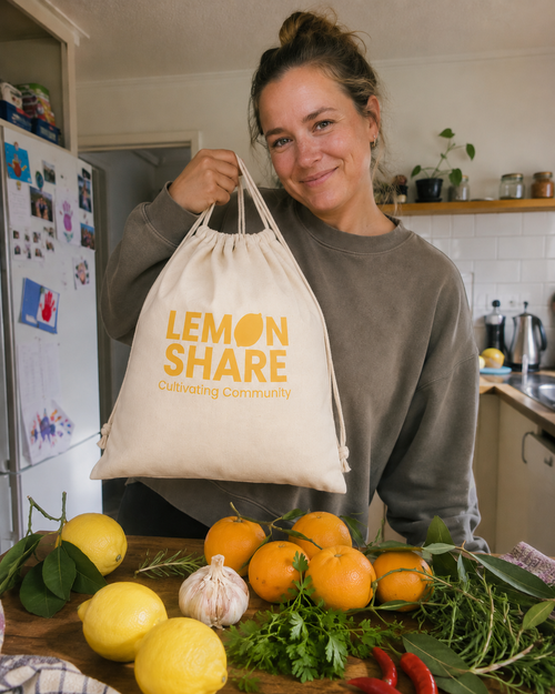 A woman in a kitchen holds up a cream-colored canvas drawstring bag with yellow text that reads 'LEMON SHARE Cultivating Community'. In the foreground, a wooden counter is filled with fresh lemons, oranges, garlic, herbs, and red chilies.