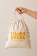 A hand holding a beige drawstring bag with yellow text that reads 'LEMON SHARE' and 'Cultivating Community' against a plain beige background.
